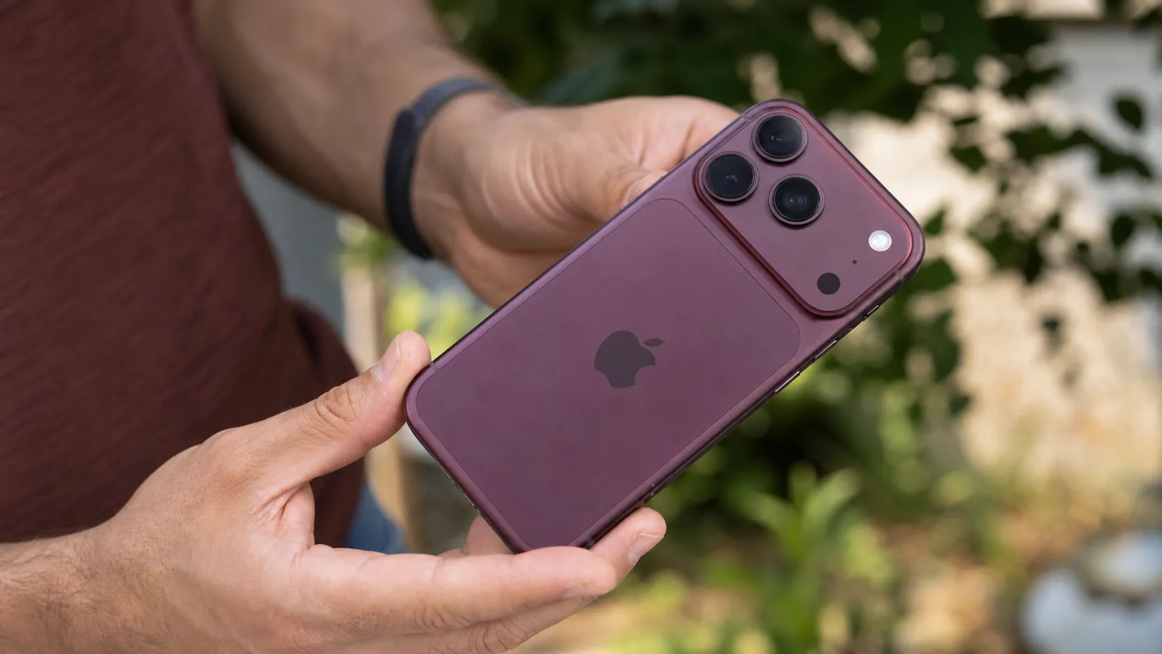 Hands holding the Burgundy iPhone 18 Pro outdoors showcasing the titanium back and camera array.